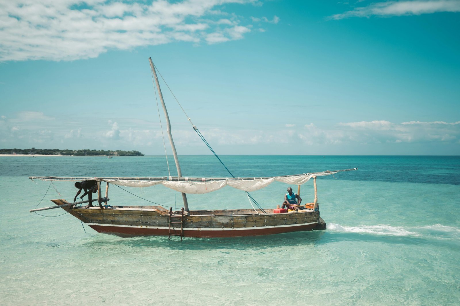 Two local fishermen navigate a traditional dhow on the clear waters of Zanzibar under a bright blue sky.